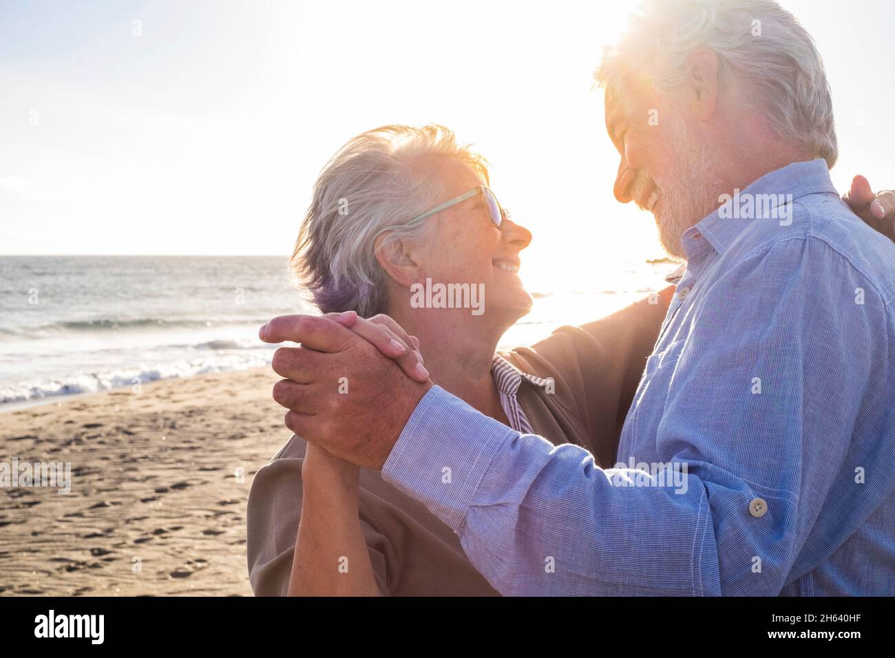 couple of two old and mature happy seniors enjoying summer dancing together at the beach on the sand with the sunset at the background. retired and leisure lifestyle Stock Photo