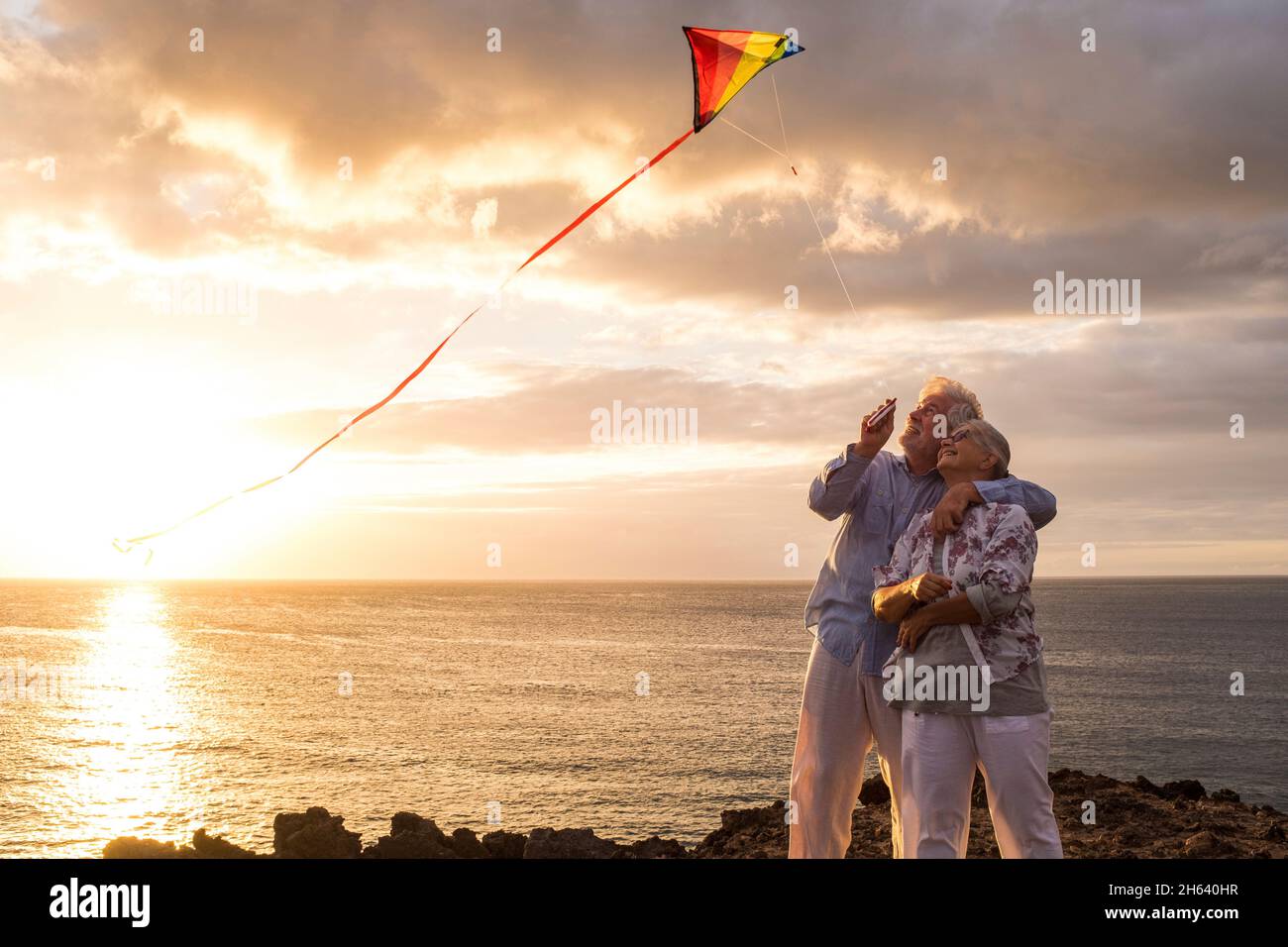 close up and portrait of two old and mature people playing and enjoying with a flaying kite at the beach with the sea at the background with sunset - active seniors having fun Stock Photo