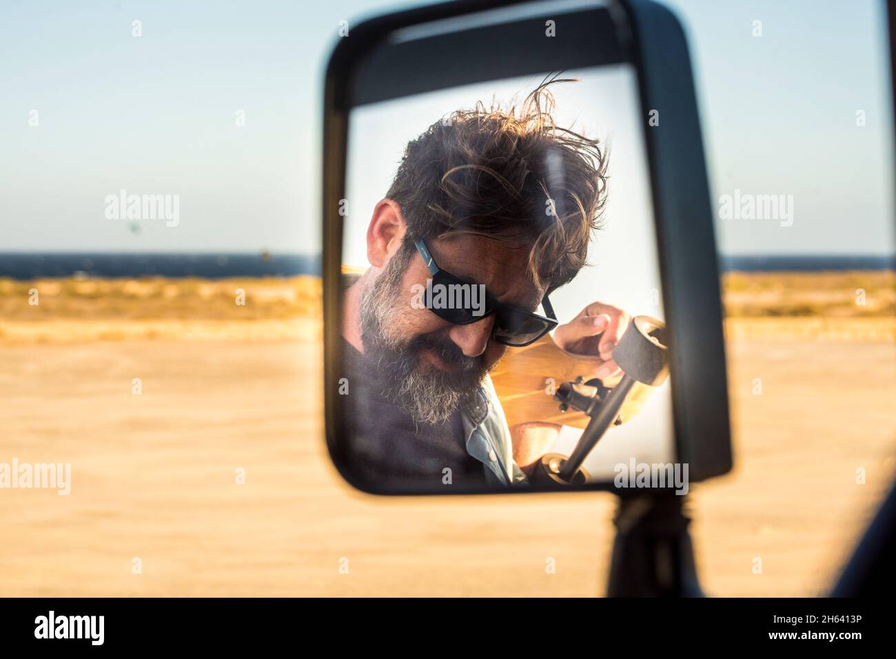 bearded handsome adult man viewed in car mirror - summer travel adventure and active lifestyle male people with yellow desert and ocean in background - concept of journey Stock Photo