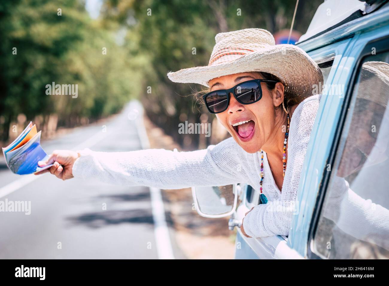 overjoyed adult pretty woman outside the window of her classic car with guida map on hand asking help or have fun - long road in background for travel adventure lifestyle people concept Stock Photo