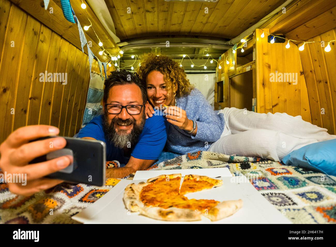 adult couple eat pizza and take selfie inside old clasic vintage home made wooden van in travel vacation together smiling and having fun - alternative adventure home camper lifestyle people Stock Photo