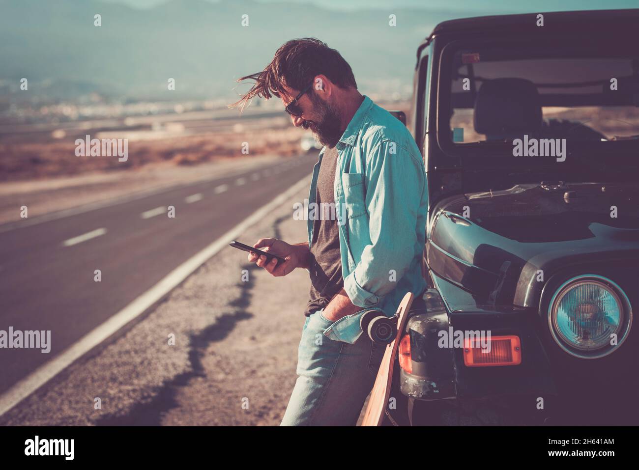 young handsome man use phone cellular outside the black car vehicle with long road in background - travel people adventure concept with roaming internet wireless connection technology Stock Photo