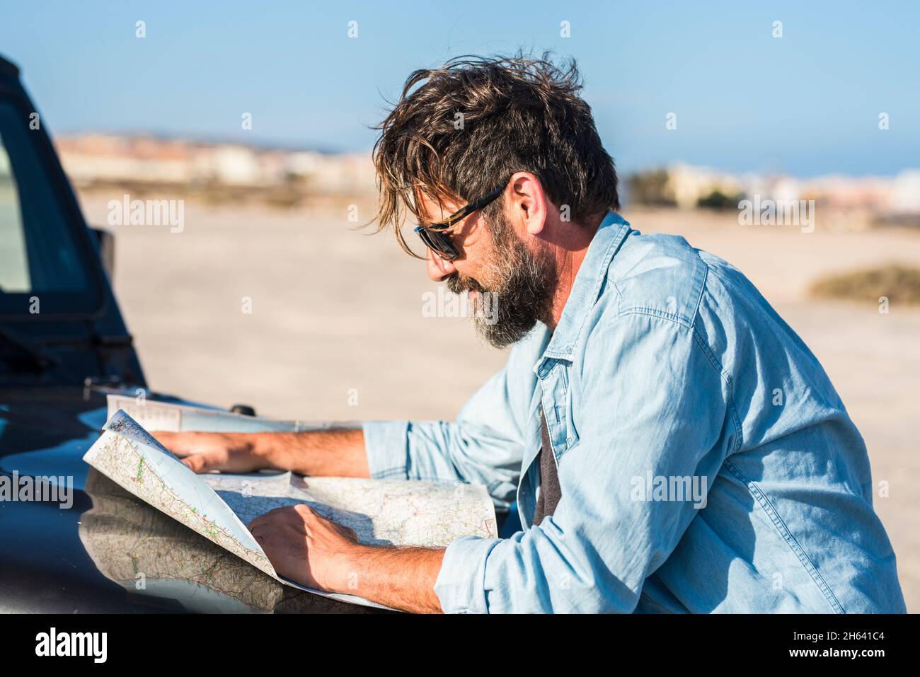 man look a paper road trip map standing outside the car with blue sky and ground desert in background - concept of travel and adventure lifestyle people - outdoor leisure travel vacation activity Stock Photo