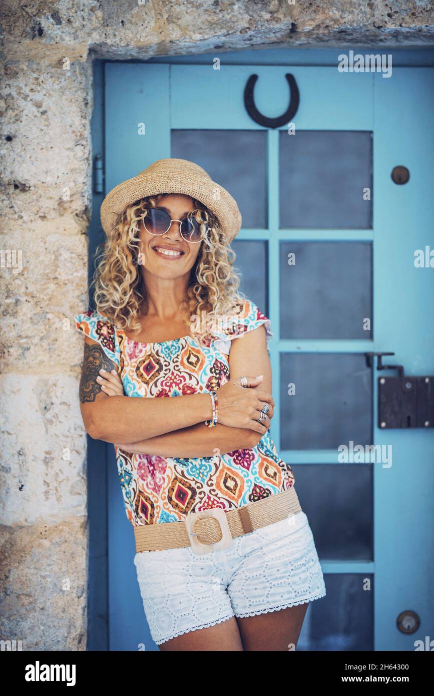 portrait of beautiful smiling hipster young woman in sunglasses and straw hat leaning on wall with her arms crossed in front of a closed door. pretty tattooed woman posing with her arms folded. Stock Photo