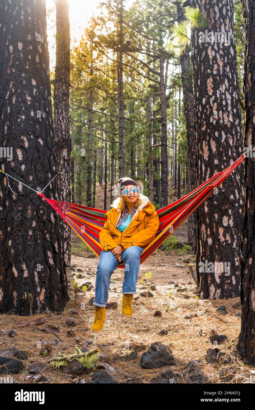 woman with yellow jacket sit down and rest on a colorful hammock on the forest woods enjoying outdoor leisure activity and adventure weekend or vacation time - people and nature Stock Photo
