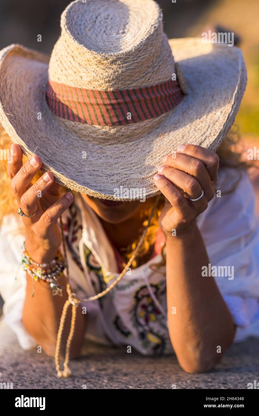 woman holding large hat with hands and obscured face looking down. styllish woman with bracelet holding large straw hat looking down and leaning on her elbows Stock Photo
