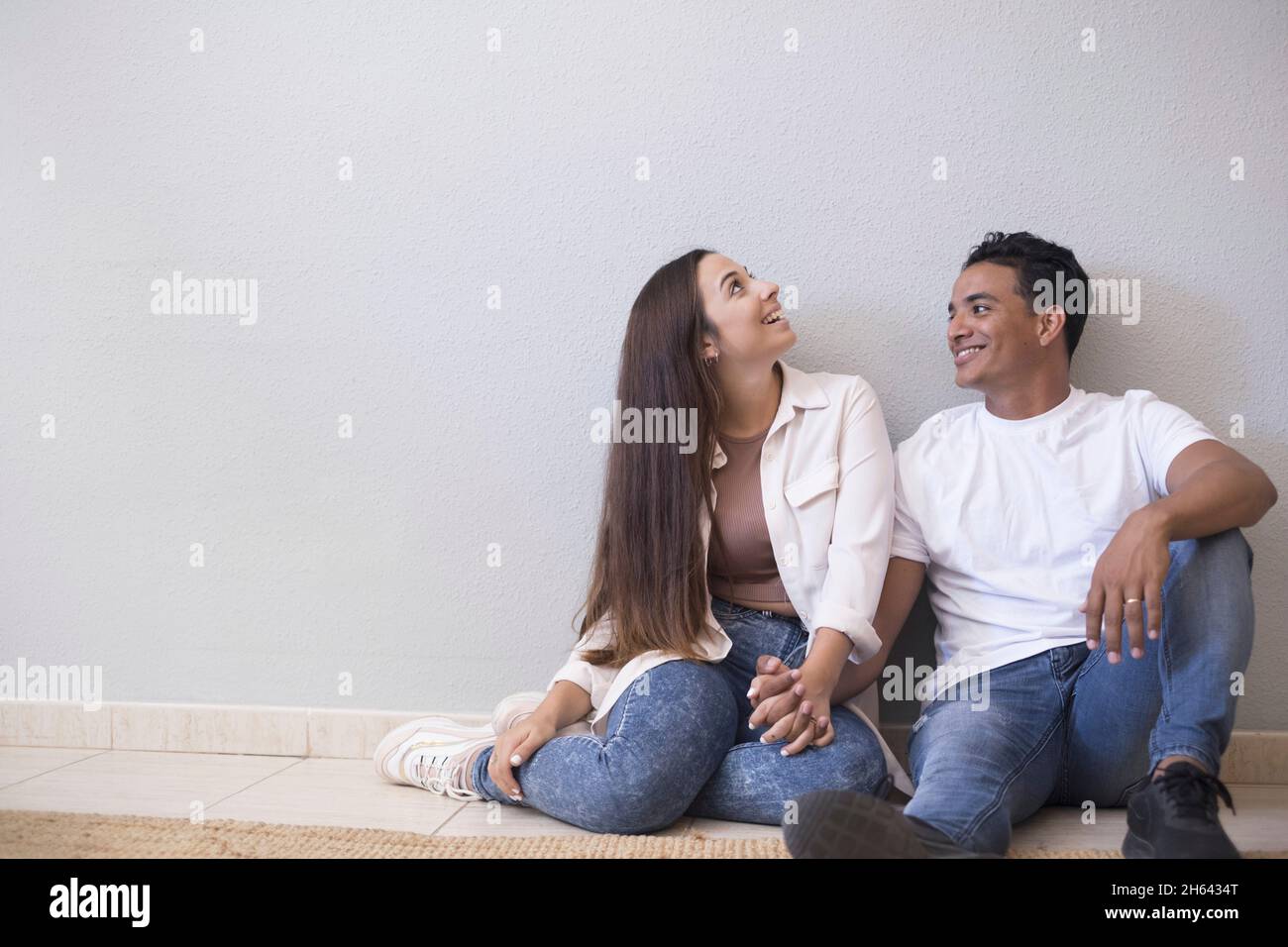 young couple sitting on floor of living room holding hands and leaning on wall. couple spending quality time at home. cheerful couple sitting on the floor of new house Stock Photo