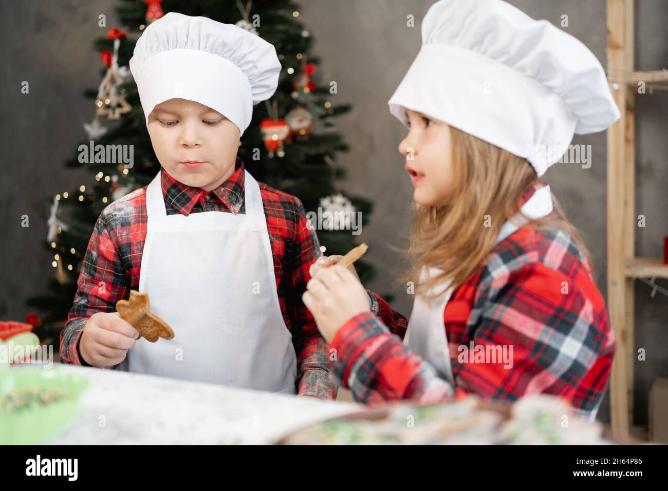 Happy little kids eating Christmas cookies in the kitchen, brother and sister in chef uniform are cooking at home, kids and Christmas gingerbread. Stock Photo