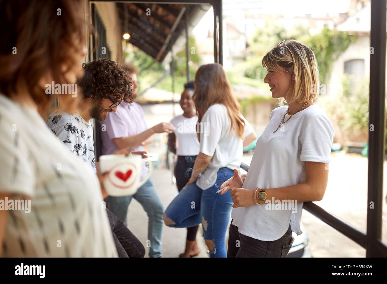 group of young adult people having a break at work, standing outdoor, smoking cigarettes, drinking coffee, talking, smiling Stock Photo