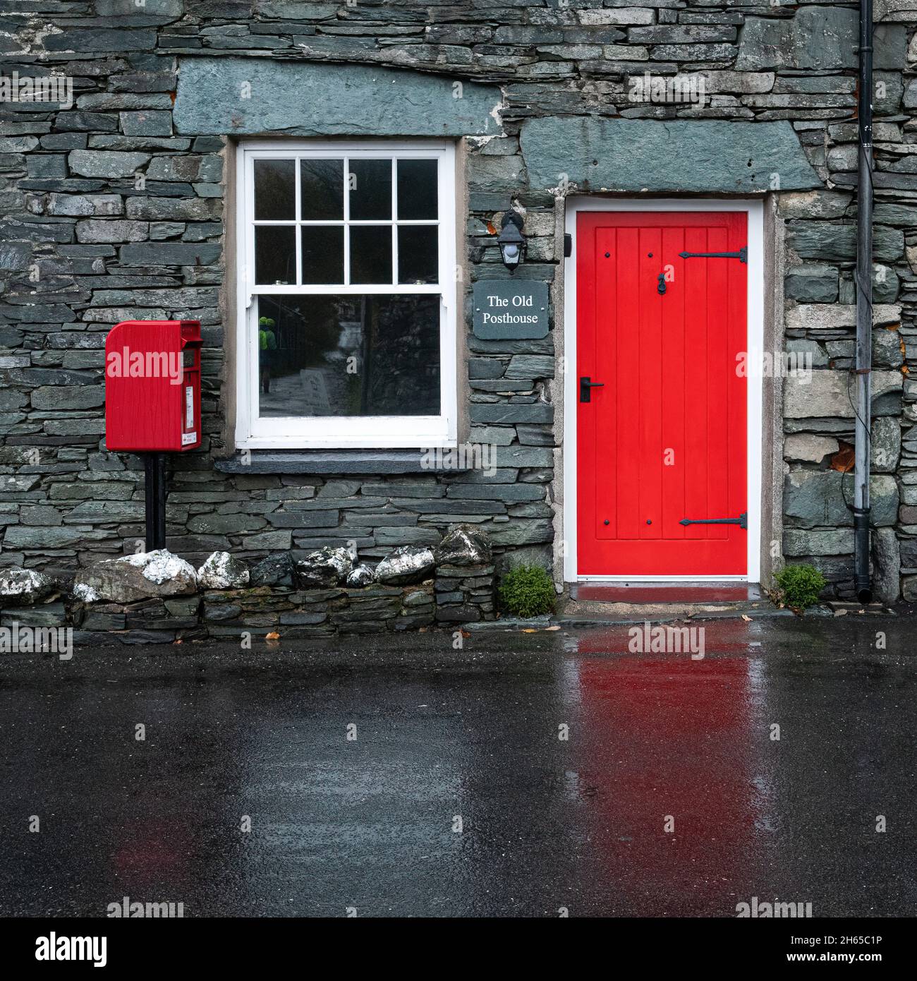 The Old Posthouse in the pretty Lake District village of Rosthwaite, Cumbria, England, UK, with a red door reflected on the wet road Stock Photo