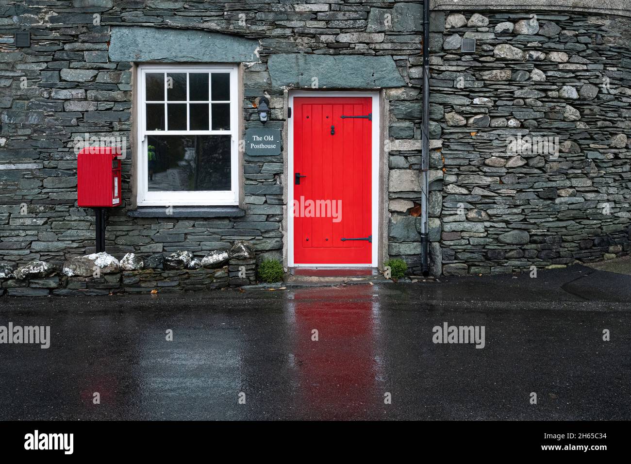 The Old Posthouse in the pretty Lake District village of Rosthwaite, Cumbria, England, UK, with a red door reflected on the wet road Stock Photo