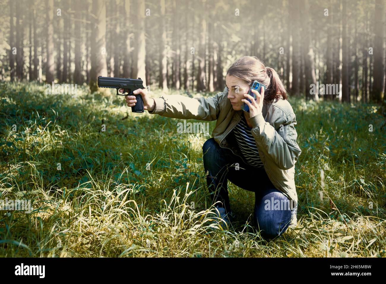Woman crouching and aiming a gun and using a mobile phone. Stock Photo