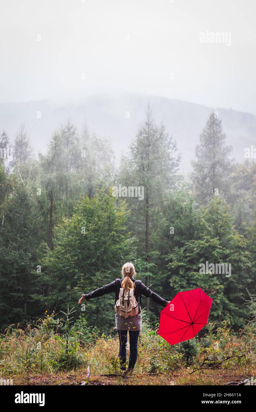 Bad weather can not ruin her happy day. Woman with red umbrella standing in rain and fog at forest. Female backpacker with arms outstretched. Feel fre Stock Photo