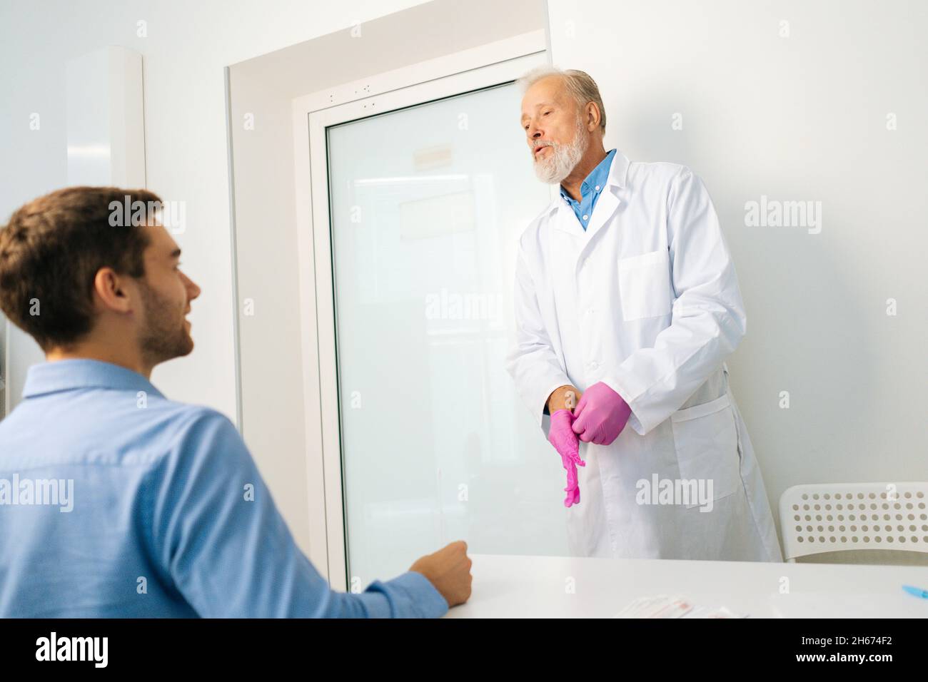 View from back of sitting male patient to mature adult urologist putting pink protective gloves in hands before examining client. Stock Photo