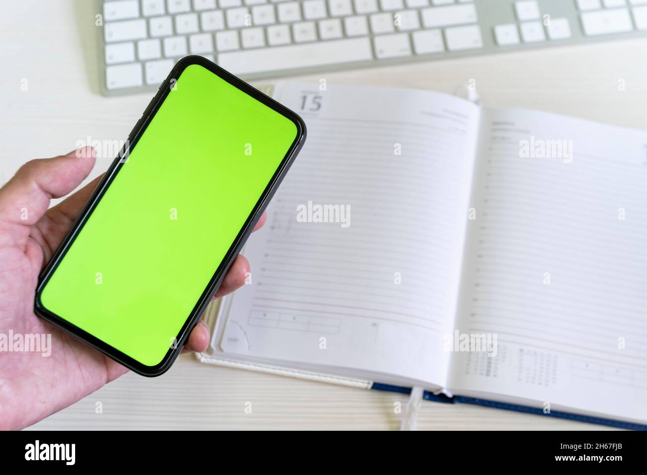 A smartphone with a green screen in a man's hand against the background of a business notepad and a computer keyboard. Stock Photo