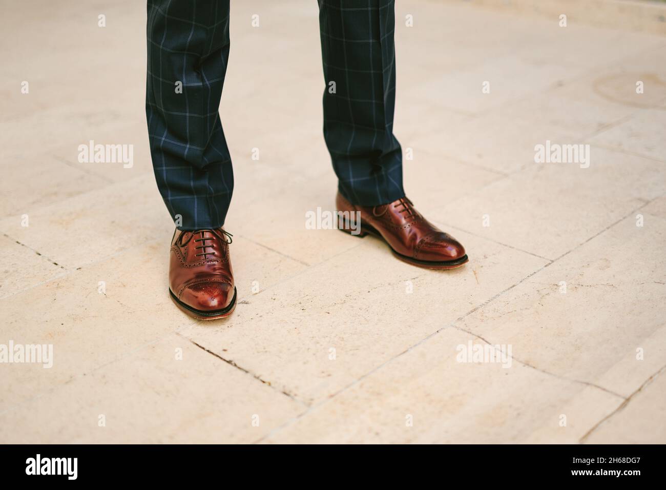 Man in black plaid trousers and brown patent leather shoes stands on a tile floor. Legs close up Stock Photo