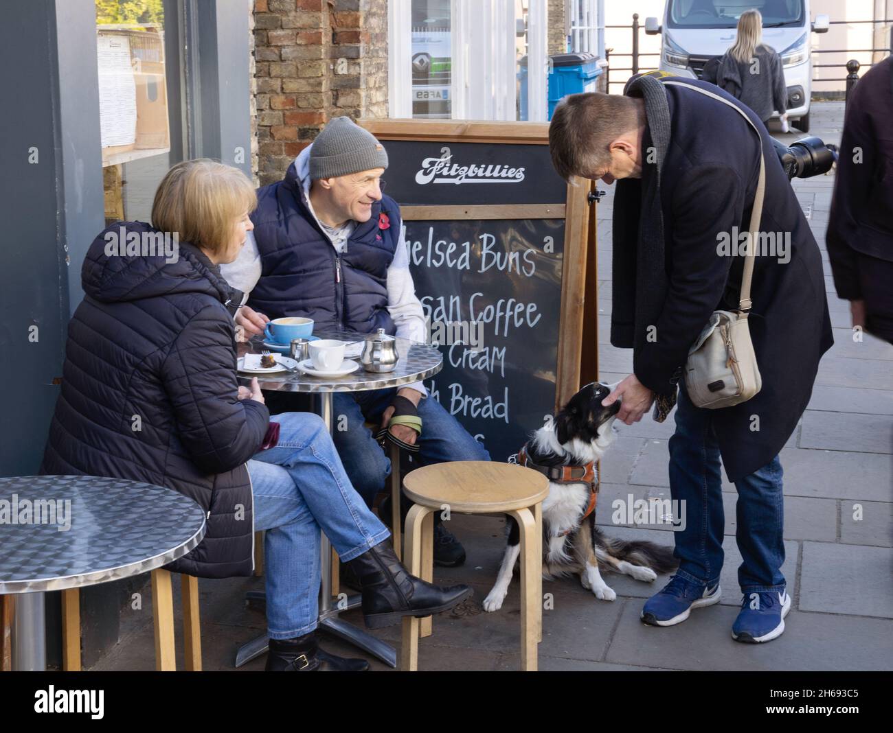 Street photography UK;  a couple having coffee with their pet dog, and man passer by petting dog - British love of dogs; Cambridge UK Stock Photo