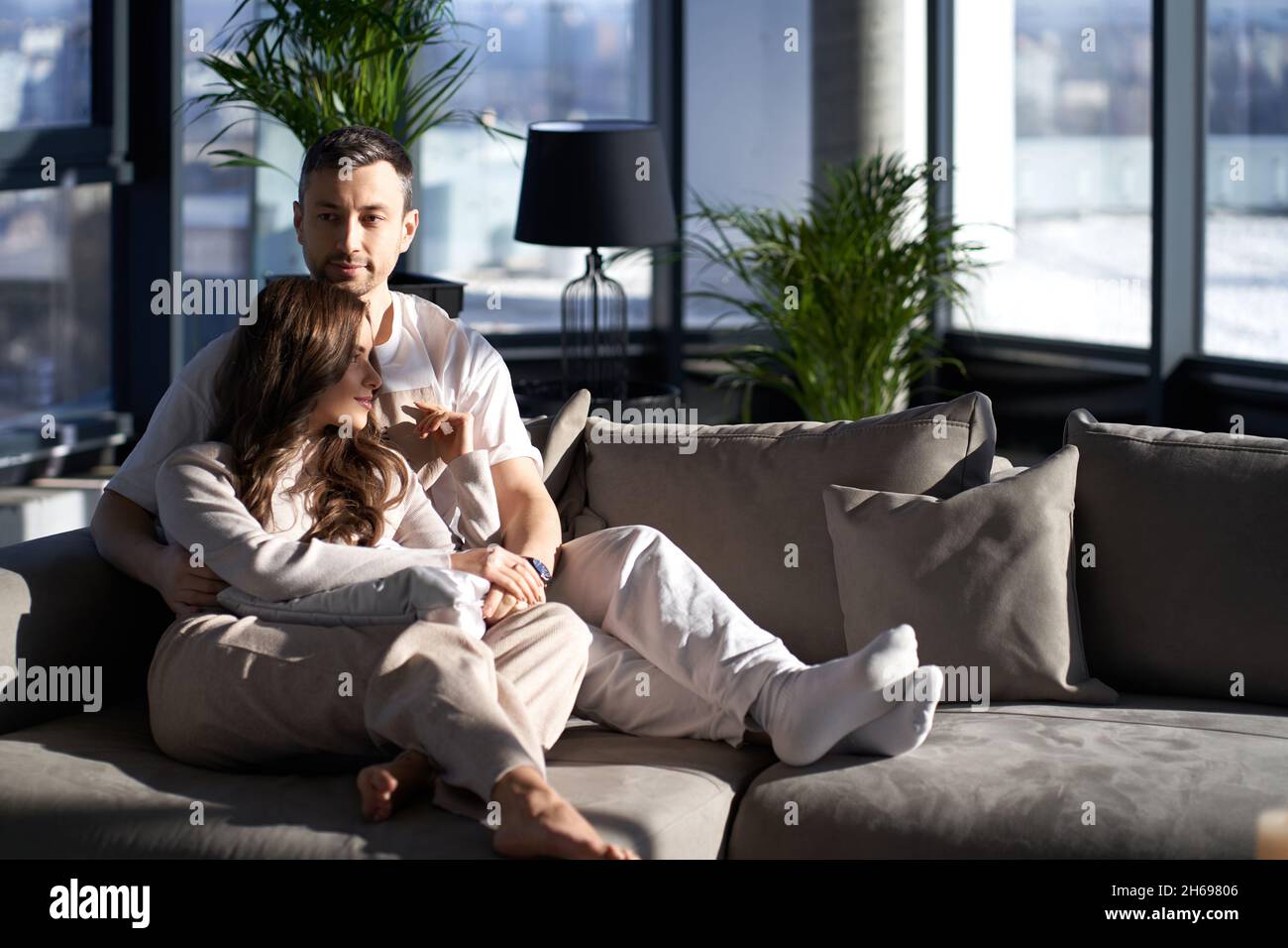 Handsome caucasian man and beautiful woman sitting in hugs while resting together on comfy couch. Daylights from panoramic windows at modern apartment. Relaxation at home.  Stock Photo