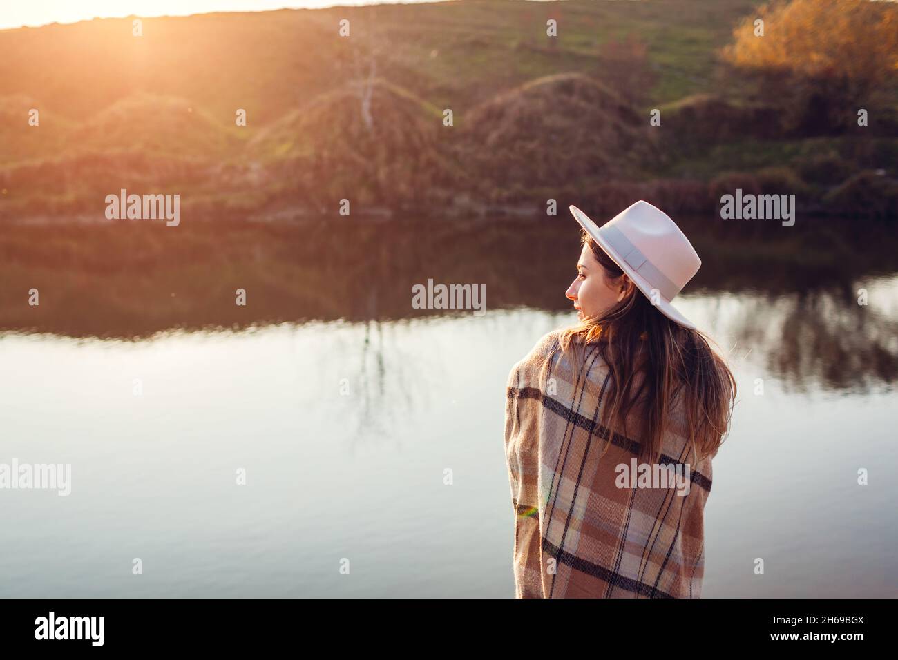 Back view of woman relaxing by autumn river at sunset. Stylish girl in hat enjoying fall landscape in silence. Harmony and balance. Healing nature Stock Photo