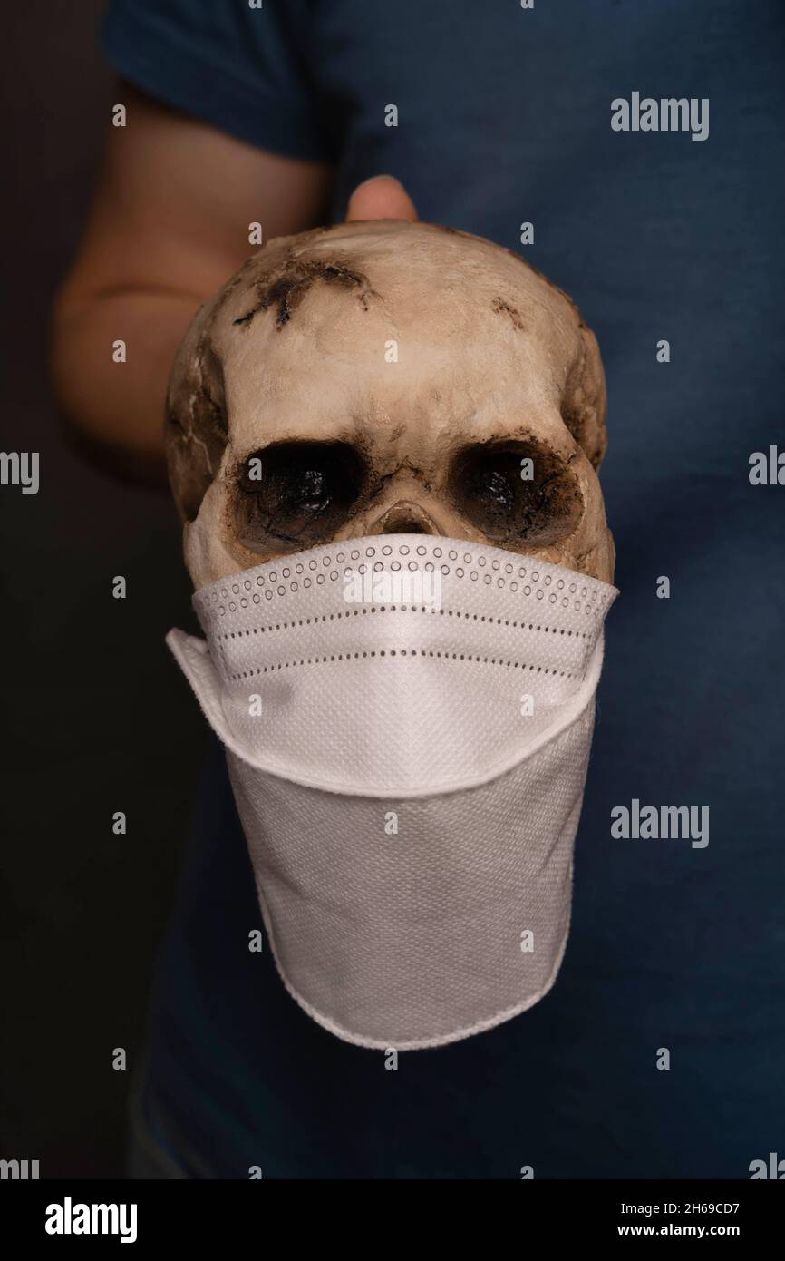 White man holding human skull that wears face mask in blue shirt on grey background Stock Photo