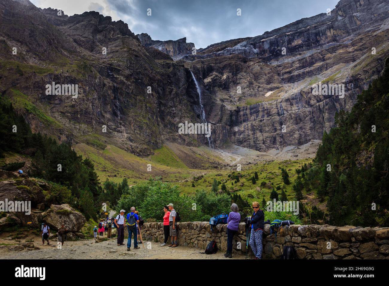 Hikers walk in the Cirque de Gavarnie in the French Pyrenees National Park, a UNESCO World Heritage Site. A large waterfall dominates the  cirque. Stock Photo