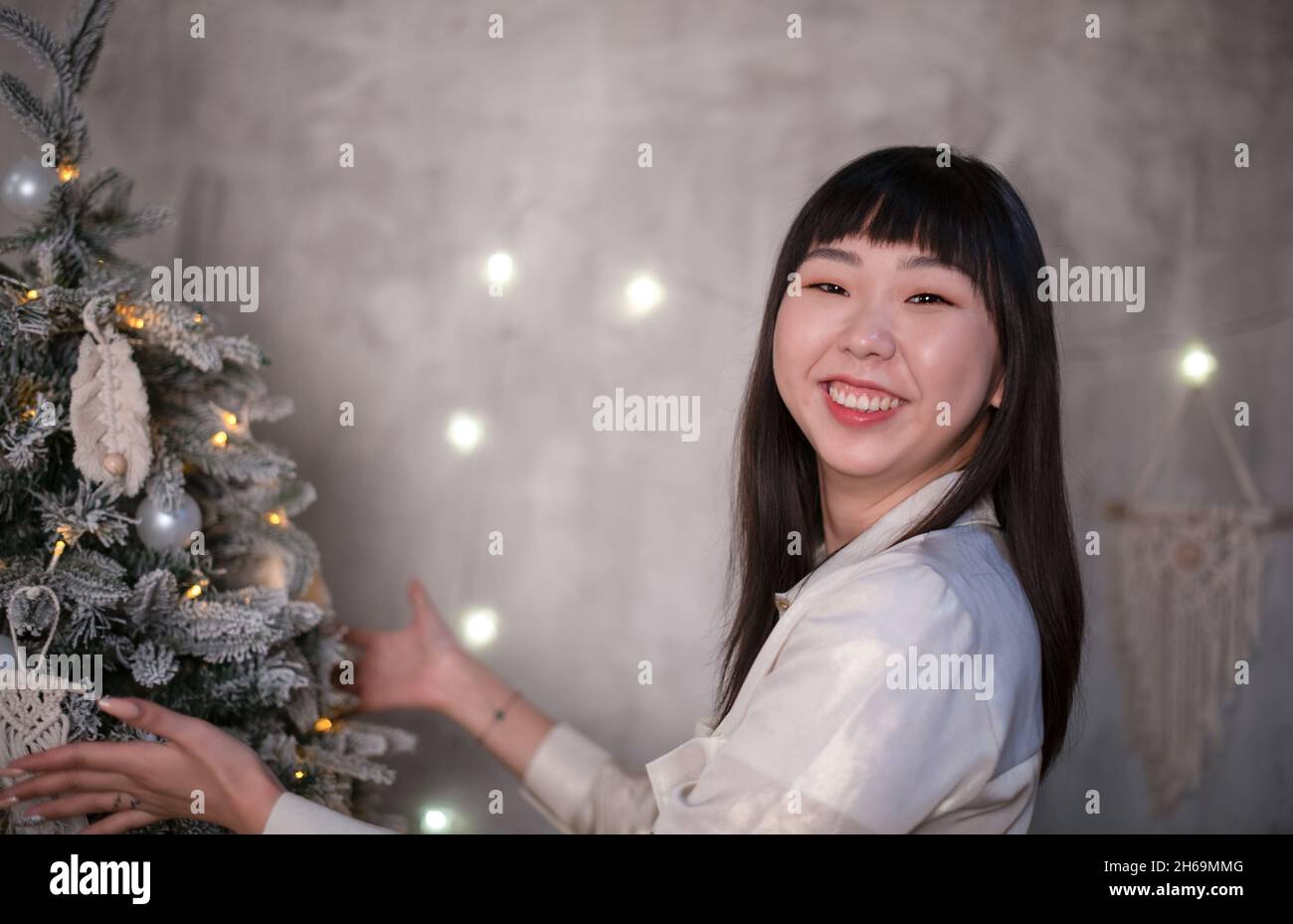 A young, beautiful Korean Asian woman dresses up a Christmas tree with toys made of natural macrame in the style of zero waste at home with positive Stock Photo