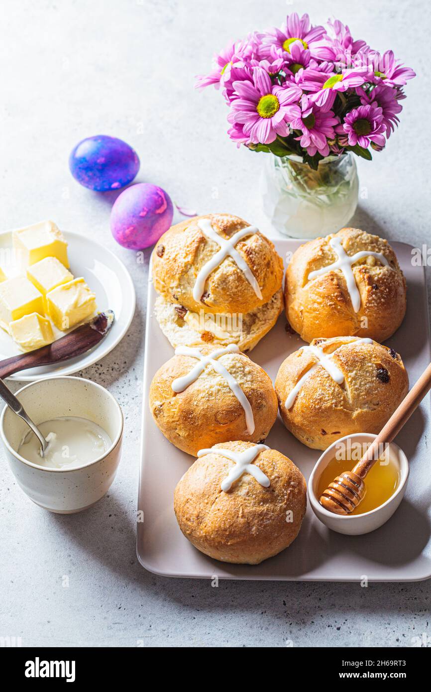 Traditional Easter cross buns with raisins, butter and honey on gray background with flowers and colored eggs. Stock Photo