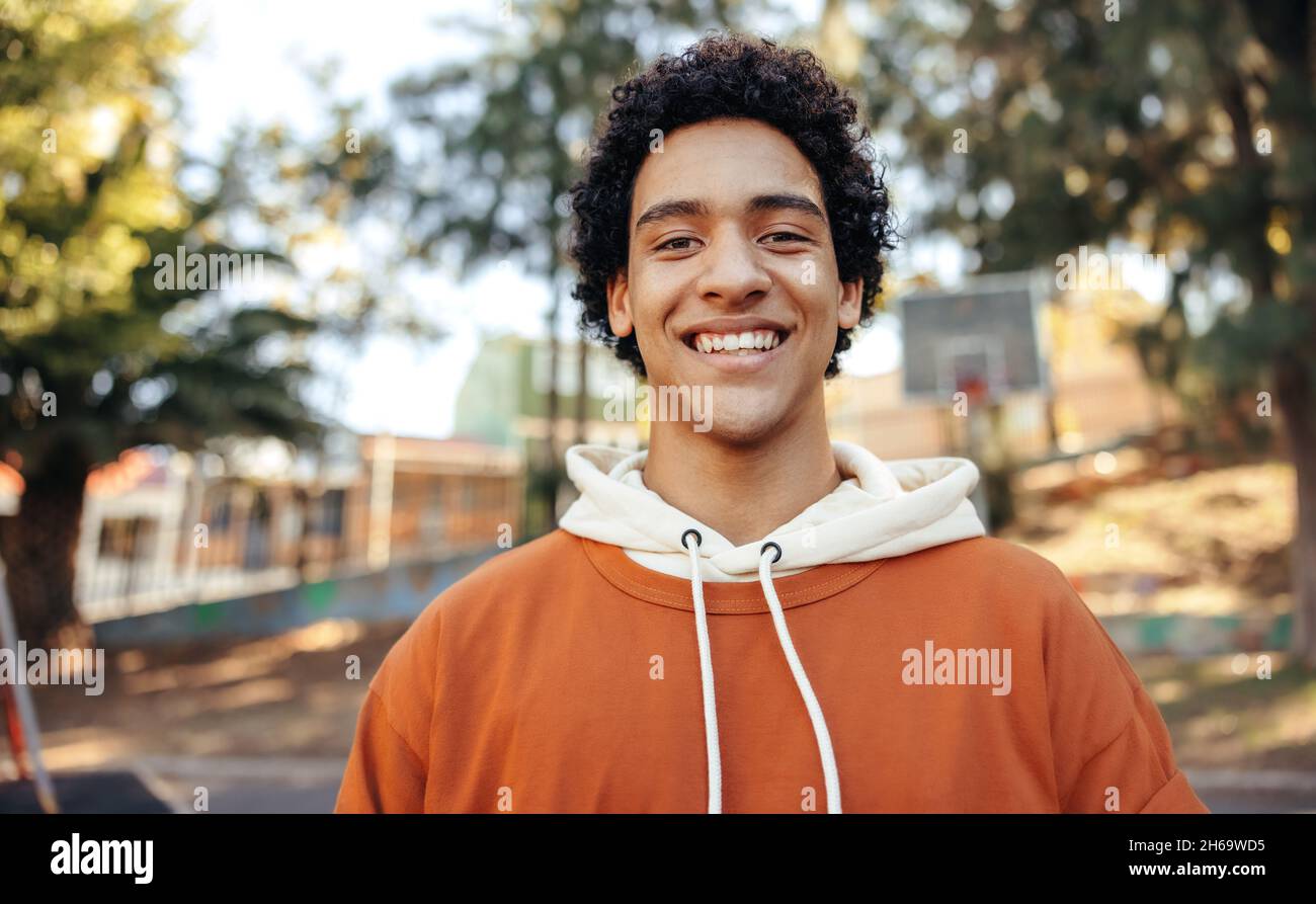 Happy male youngster smiling at the camera outdoors. Fashionable teenager wearing casual clothing in an urban park. Cheerful teenage boy standing alon Stock Photo