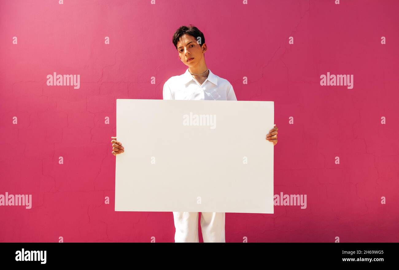 Teenage gay activist displaying a blank banner in a studio. Assertive teenage boy holding a white placard against a pink background. Young queer boy c Stock Photo