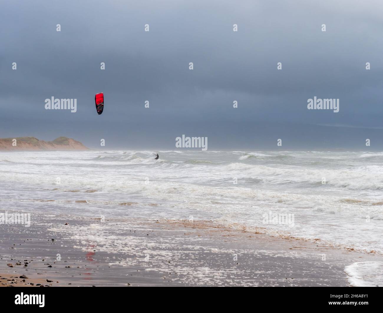 A kitesurfer surfing along the waves on the beach against a stormy, cloudy sky Stock Photo