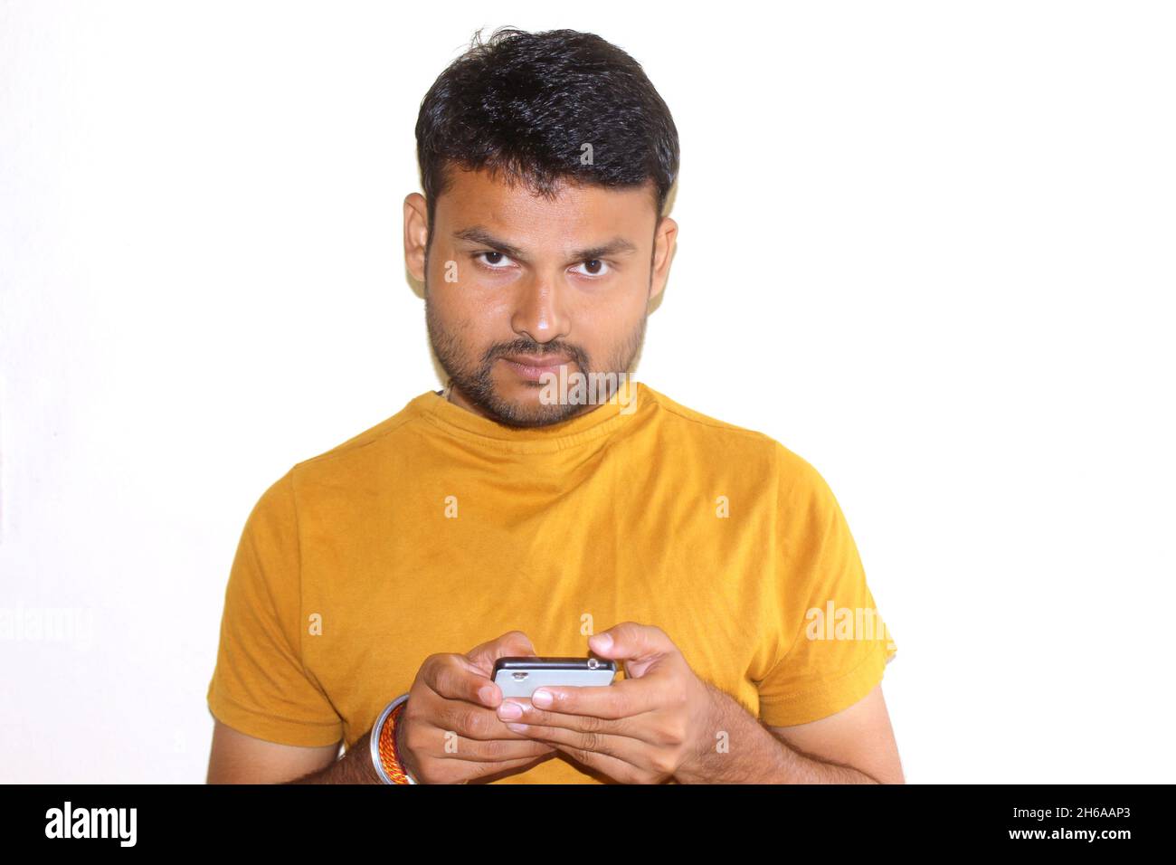 Attractive male using mobile phone, thinks about something, looks doubtfully upwards, Indian male student texting on smart phone, .against white concr Stock Photo