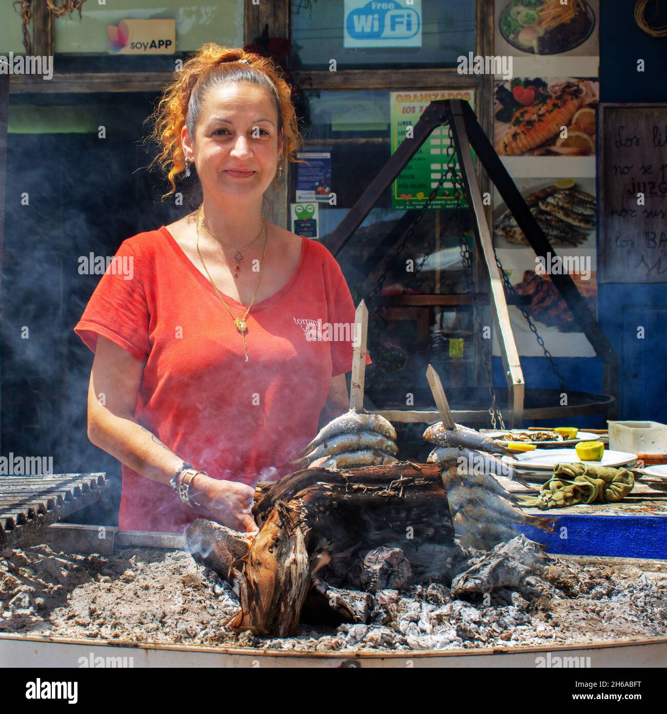 Spanish lady grilling sardines over wood fire in Mediterranean coastal bar/restaurant, Puerto de Mazarrón, Murcia, Spain. Stock Photo