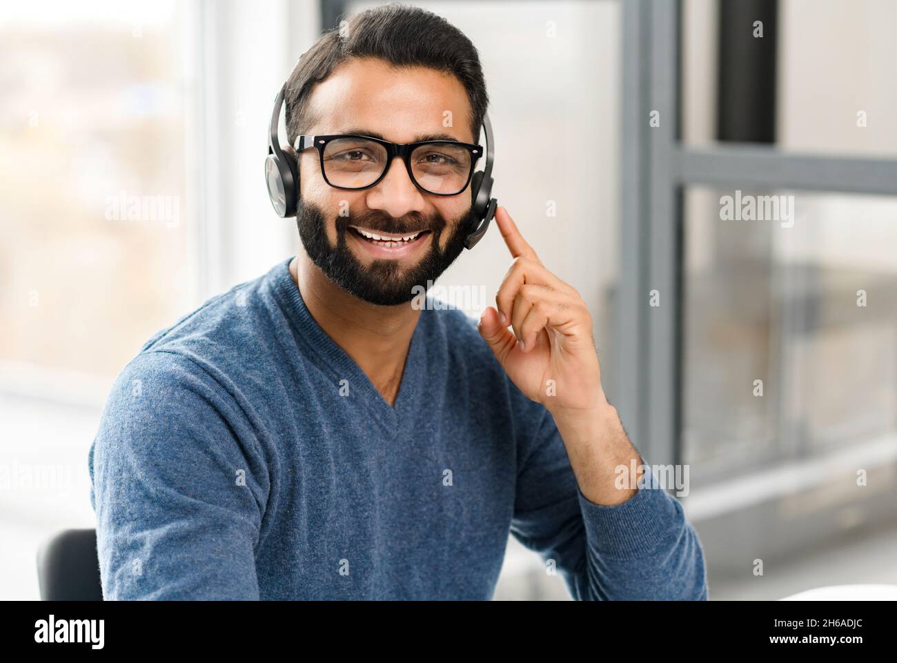 Headshot of young Indian male wearing handsfree headset and stylish glasses, in casual wear in the office, taking call and talking with customers or colleagues. Call center, help line, support service Stock Photo