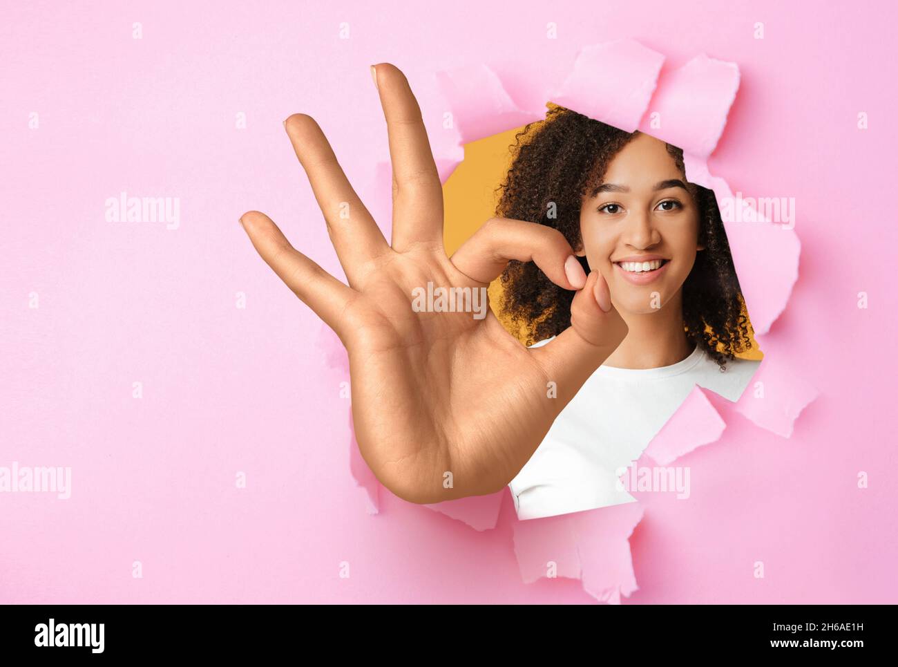 Happy young black woman showing okay gesture through hole in ripped ...