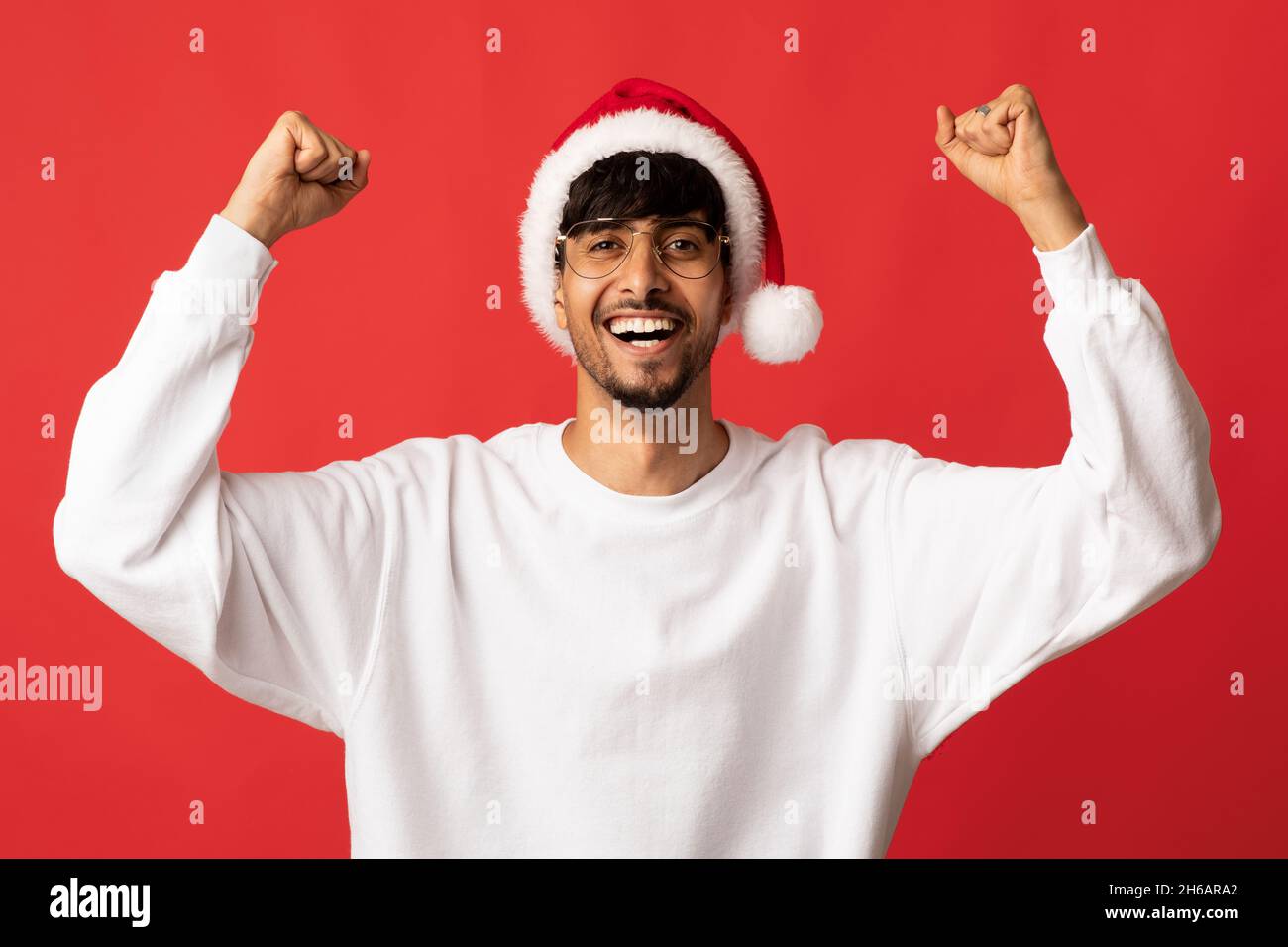 Emotional handsome millennial arabic man in red Santa hat and glasses raising hand up, having good mood, red studio background, copy space. New Year, Stock Photo