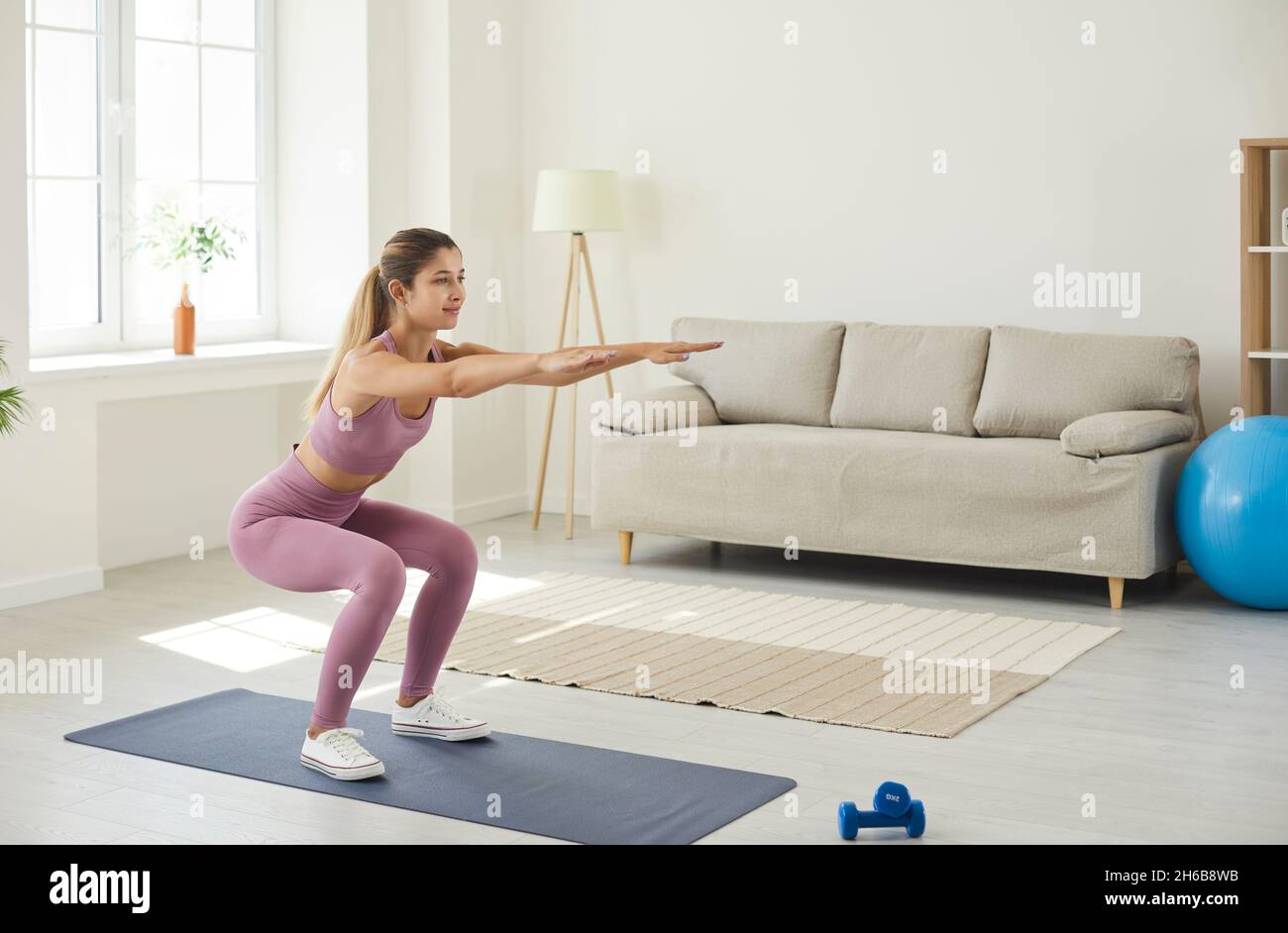 Young enduring woman trains at home doing deep squats with her arms outstretched. Stock Photo