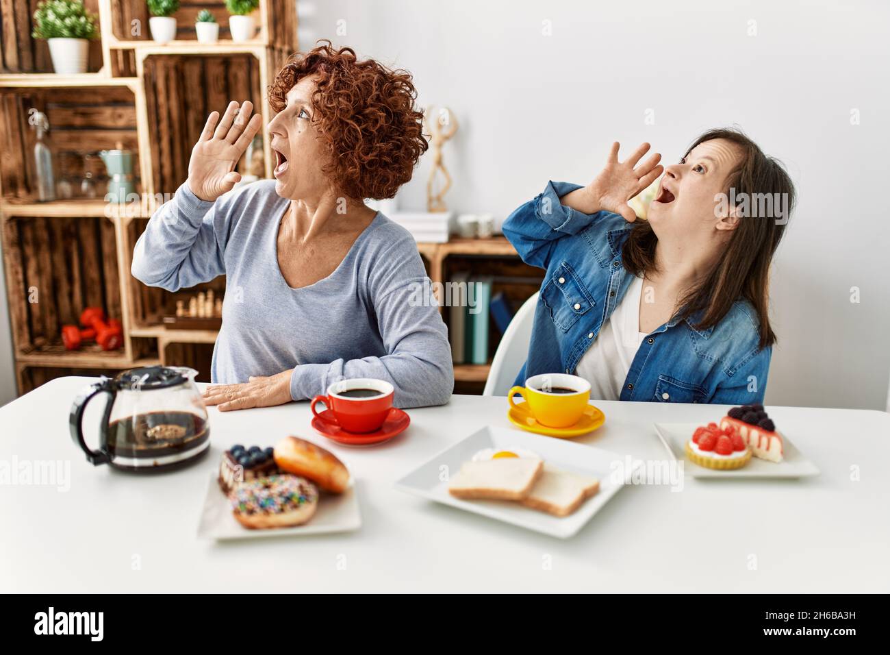 Family of mother and down syndrome daughter sitting at home eating breakfast shouting and screaming loud to side with hand on mouth. communication con Stock Photo