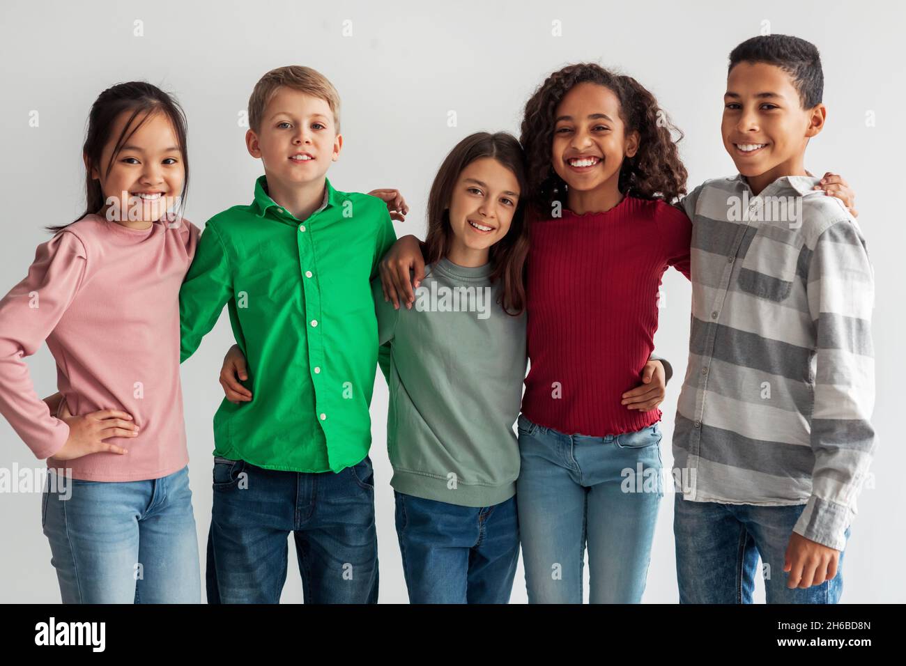 Cheerful Multicultural Preteen Kids Standing Hugging And Smiling To Camera Posing Together Over Gray Studio Background. Shot Of Mixed School Boys And Stock Photo