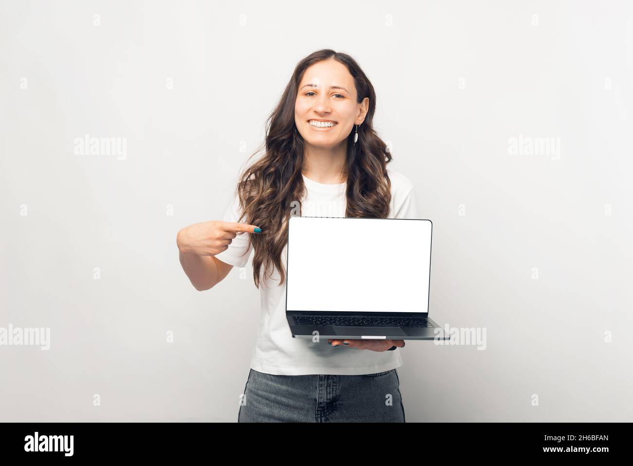 Cheerful young woman pointing at blank screen on laptop over white ...