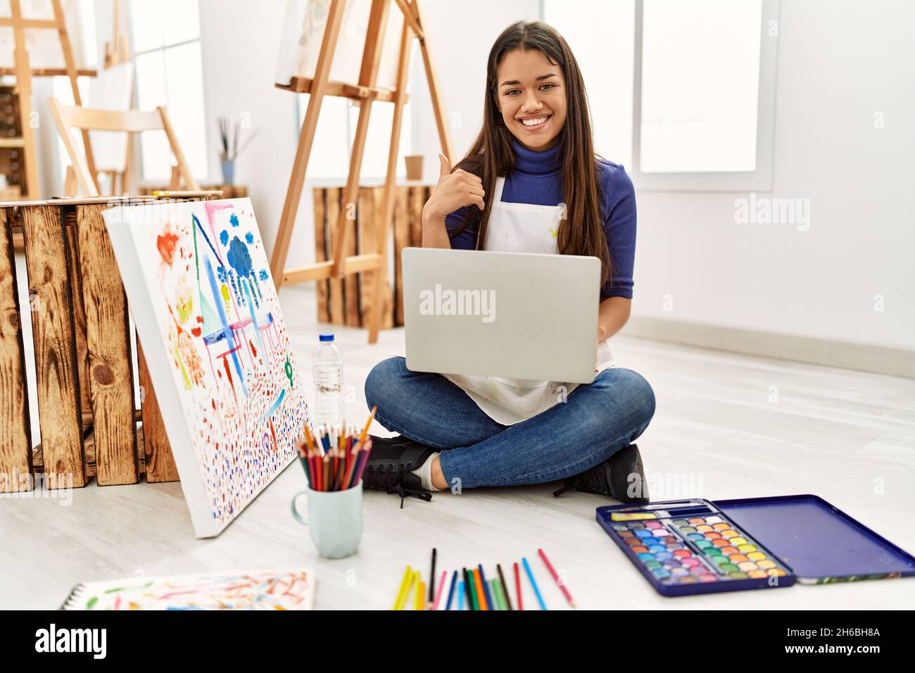Young brunette woman sitting on the floor at art studio with laptop smiling happy and positive, thumb up doing excellent and approval sign Stock Photo