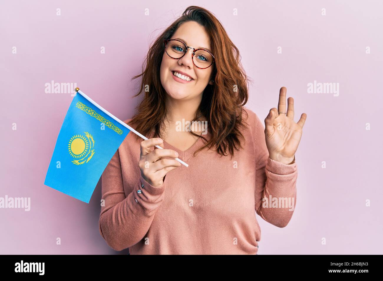 Young caucasian woman holding kazakhstan flag doing ok sign with fingers, smiling friendly gesturing excellent symbol Stock Photo