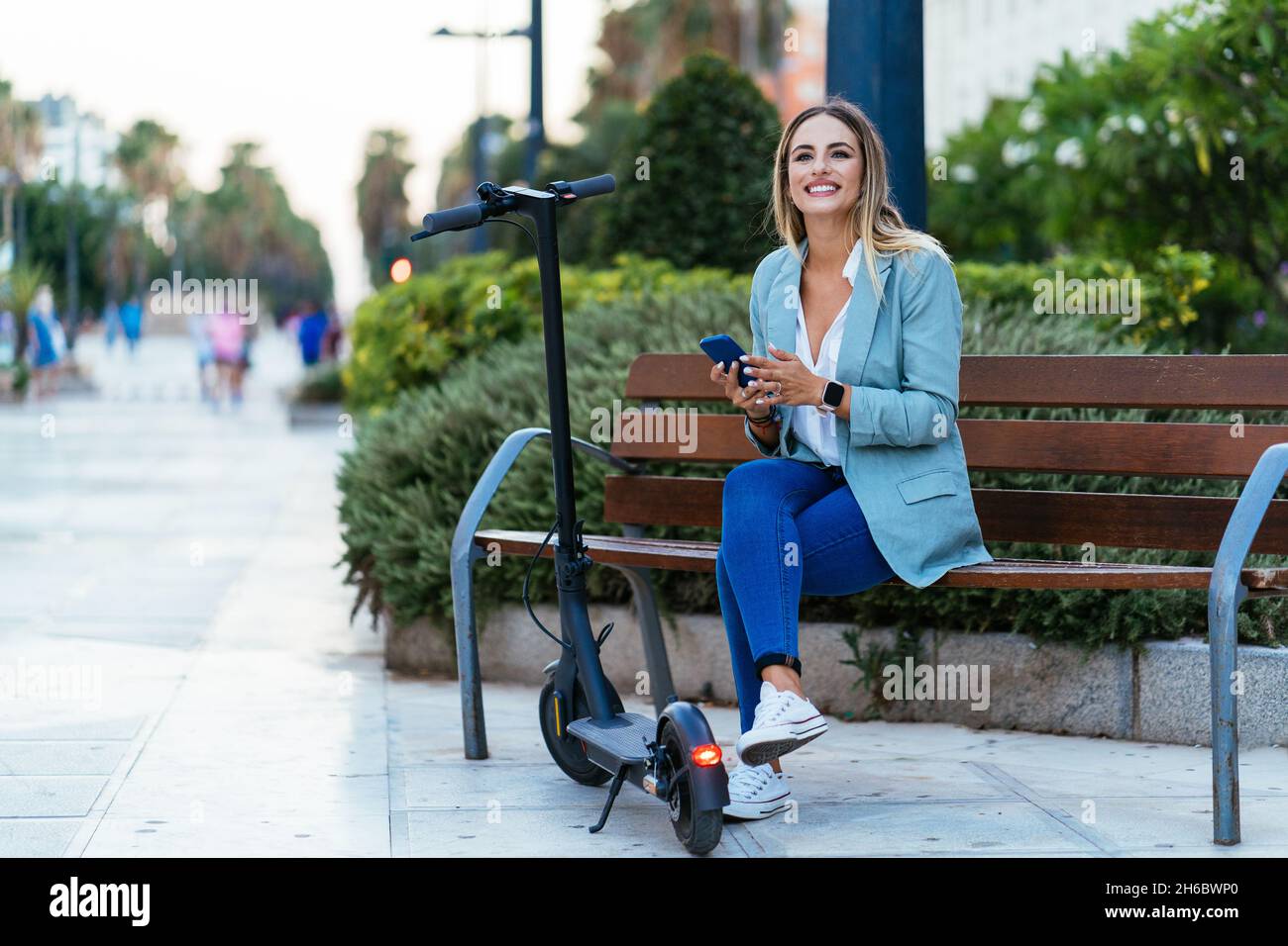 Stylish businesswoman using smartphone on bench near electric scooter Stock Photo