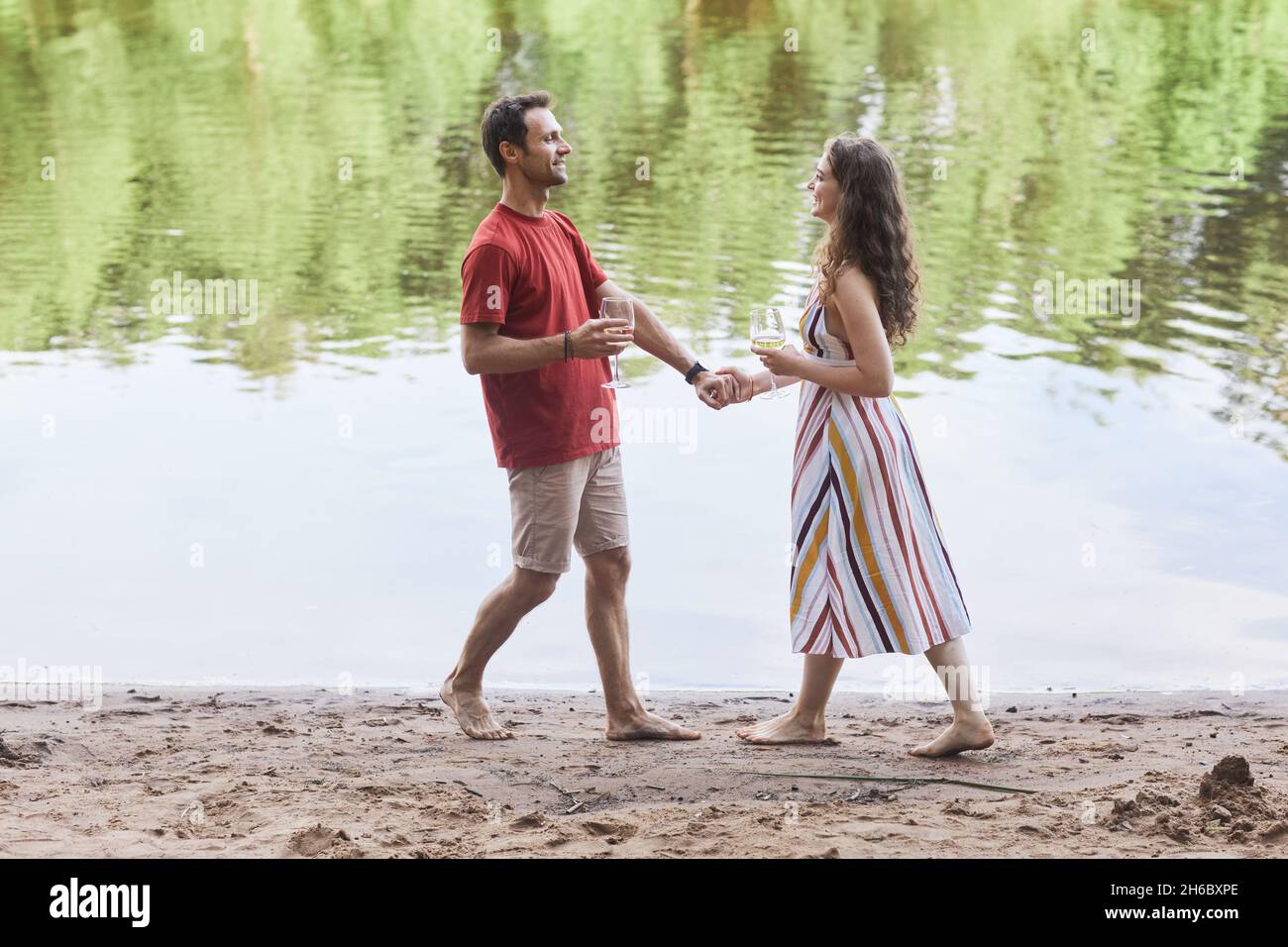 Full length portrait of happy couple holding wine glasses while enjoying romantic date by water outdoors, copy space Stock Photo