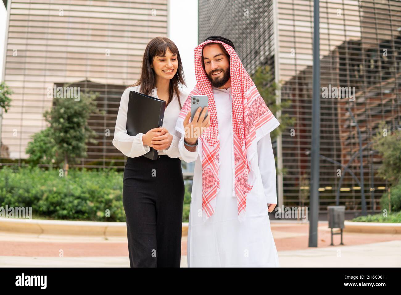 Low angle of cheerful businesswoman in Arab man in keffiyeh smiling and using smartphone together while having business meeting on street of modern city Stock Photo