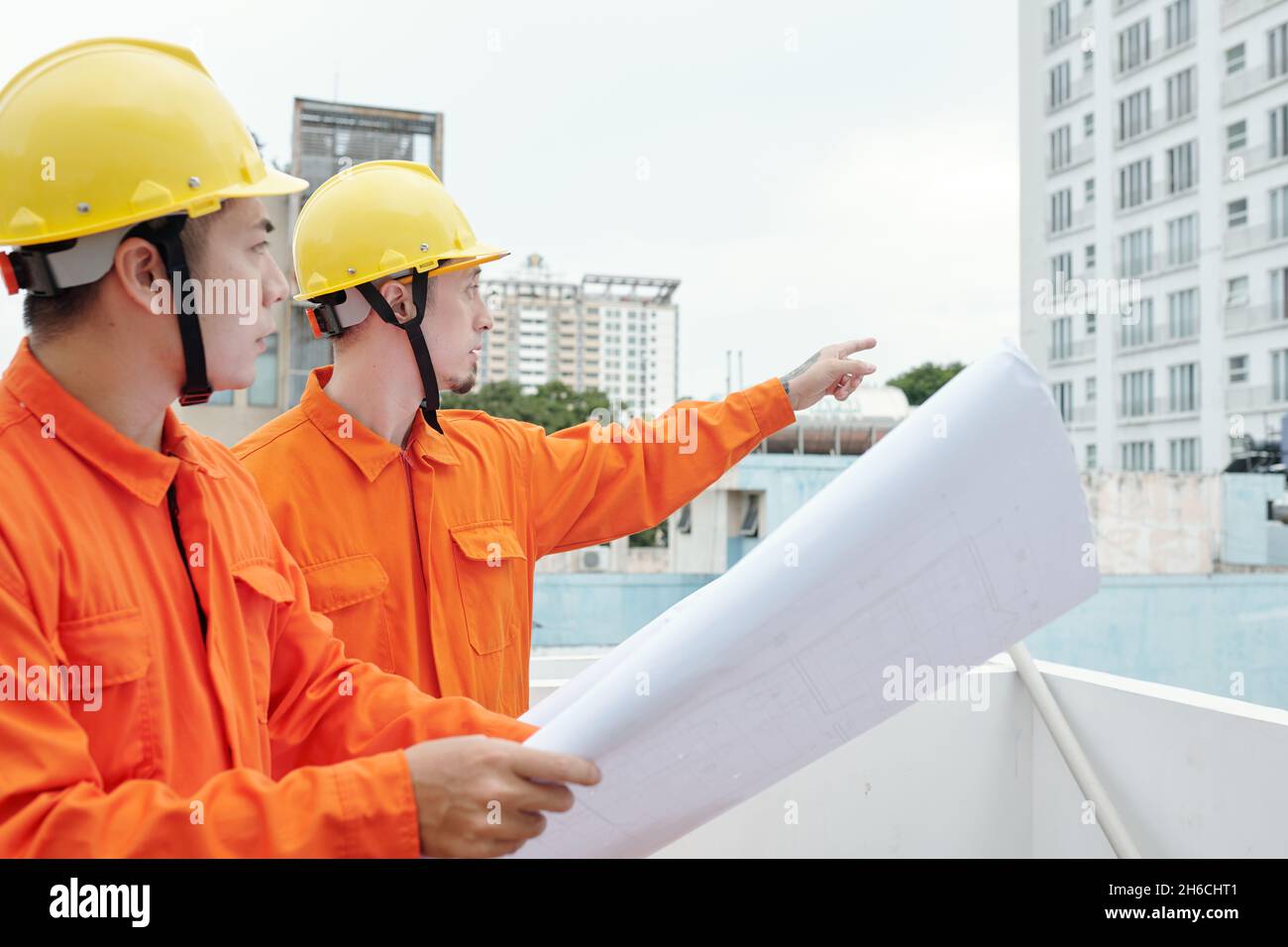 Construction workers standing at site with blueprint in hands and pointing at skyscraper near by Stock Photo