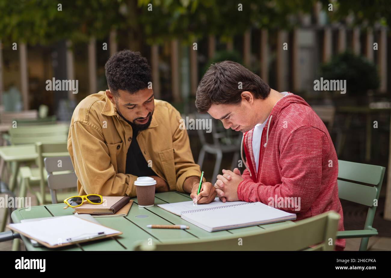 Young man with Down syndrome with his mentoring friend sitting outdoors in cafe and studying. Stock Photo