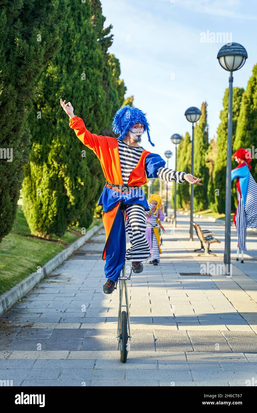 Full body of funny showman in clown costume and blue wig riding unicycle on paved pathway in park while performing show with anonymous colleagues Stock Photo