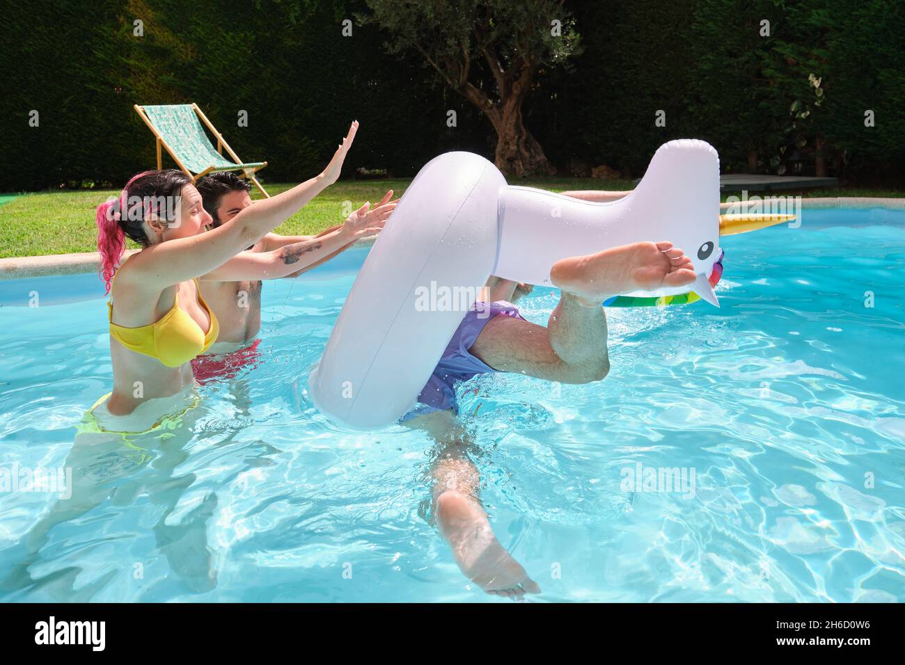 Two friends throwing man from a rainbow unicorn float into the pool. Stock Photo