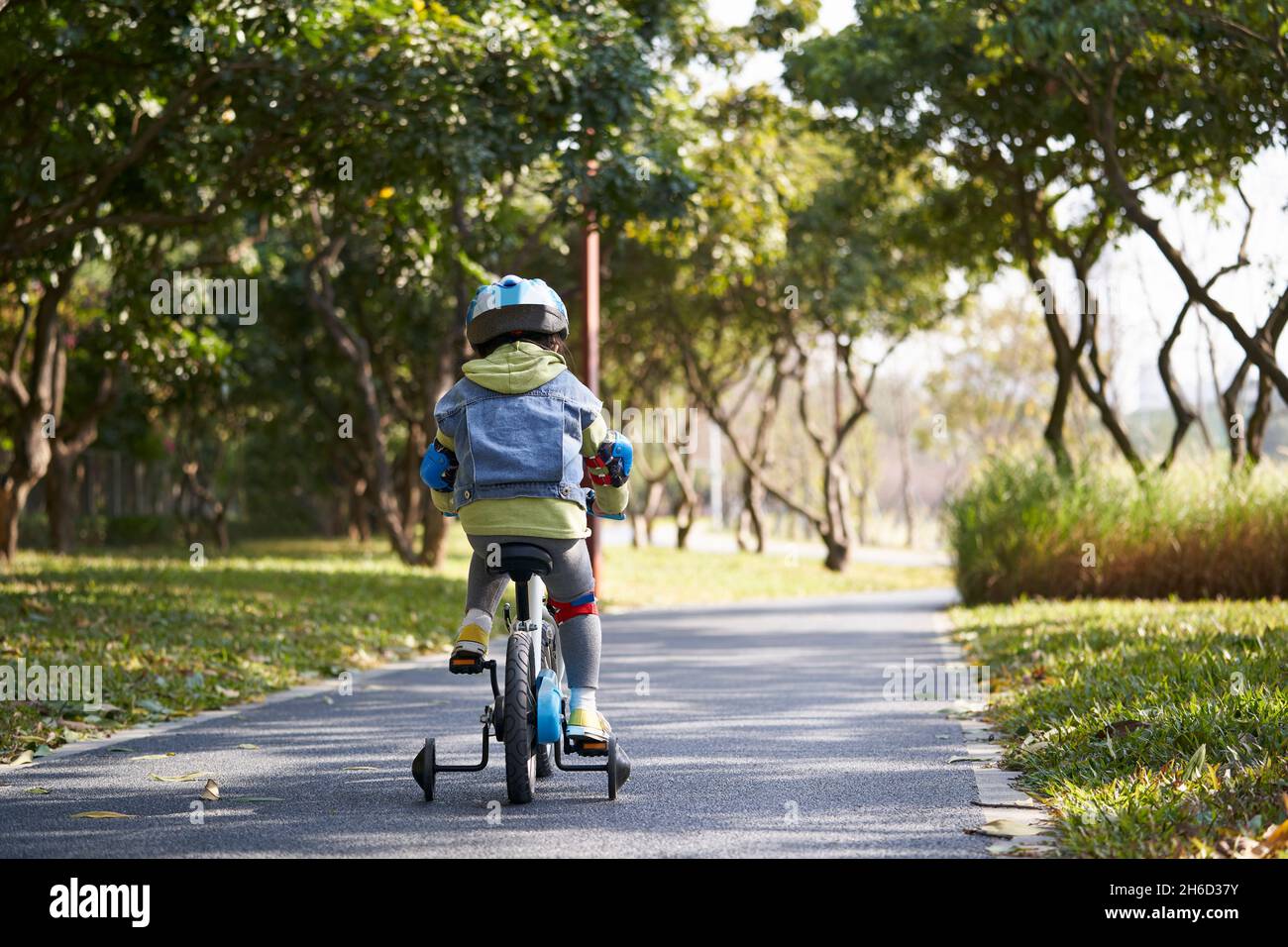 rear view of a little asian girl in full protection gear riding bike outdoors in city park Stock Photo
