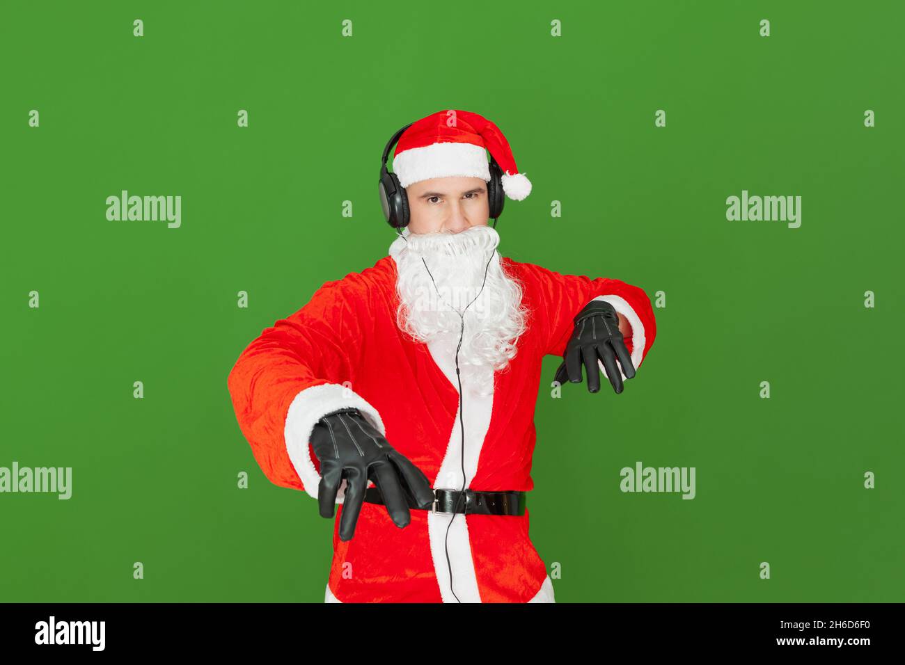 An adult Caucasian man dressed as Santa Claus with headphones is imitating a disc jockey pretending to move the records and volume control in the air. Stock Photo